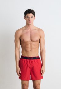 Shorts de bain rouges avec une ceinture élastique noire portant le nom de la marque "HUGO" en blanc, affichés sur un mannequin masculin torse nu.