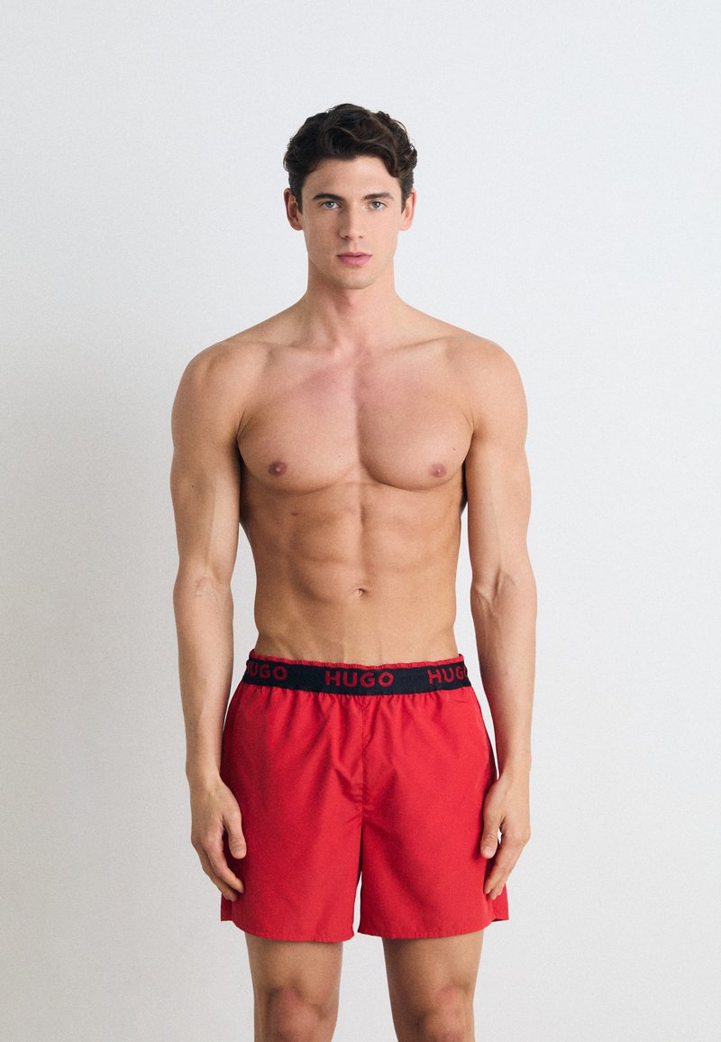 Calções de banho vermelhos com uma cintura elástica preta onde está exibido o nome da marca "HUGO" em branco, apresentados em um modelo masculino sem camiseta.