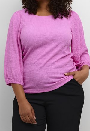 Vrouw die een gestructureerde lavendelkleurige top met lange mouwen en een zwarte broek draagt, met één hand in de zak en zichtbaar krullend haar.