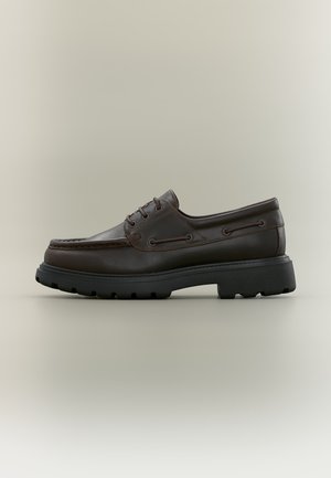 Chaussure bateau en cuir marron foncé avec semelle crantée noire épaisse et design à lacets, vue de profil sur un fond clair uni.
