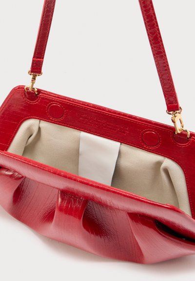 Κόκκινη τσάντα χειρός με υφή κροκοδείλου και χρυσά κούμπωμα, ανοιχτή για να αποκαλύψει το εσωτερικό από μπεζ ύφασμα και μια κεντρική λευκή ετικέτα στο εσωτερικό.