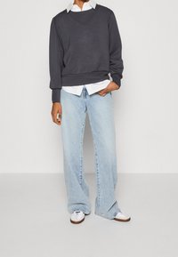 Donkergrijze sweatshirt met een V-hals, gedragen over een witte blouse met kraag, gecombineerd met lichtblauwe wijde spijkerbroek en witte sneakers.