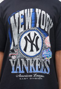 T-shirt preta de algodão com texto ousado em cinza e azul "NEW YORK YANKEES", um logótipo circular da equipa e outros logótipos e detalhes coloridos.