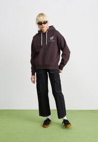 Mørkebrun jakke med glidelås og to frontlommer, kombinert med svarte vide cargo-bukser med sidelommer og hvite sømmer.