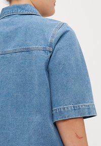 Light blue denim short-sleeve shirt showing a collar, with a plain texture and visible stitching details; a red tattoo is on the arm.