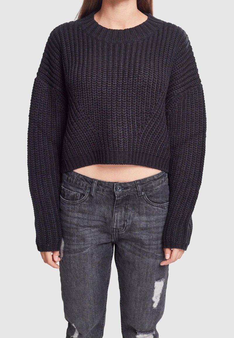 Femme portant un pull court en tricot épais noir et un jean noir délavé à taille haute avec des déchirures, posant devant un fond uni.