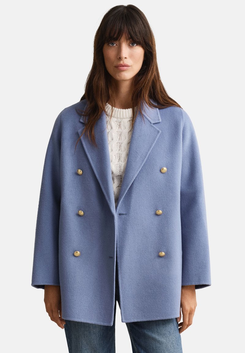 Cappotto doppiopetto in tessuto morbido azzurro chiaro con bottoni dorati. Presenta un rever a lancia e una silhouette squadrata. Indossato sopra un maglione bianco lavorato a maglia.