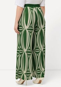 Grüne und cremefarbene weit geschnittene Hose mit geometrischem Muster, ausgestattet mit einem elastischen Bund und einer glatten Textur. Hohe Schuhe sind sichtbar.