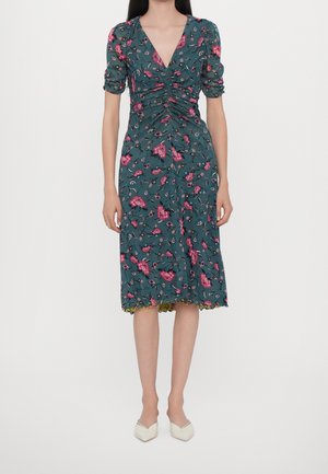 Mujer con vestido midi azul verdoso con estampado floral rosa, frente fruncido, mangas abullonadas hasta el codo y zapatos blancos puntiagudos, de pie sobre un fondo blanco.