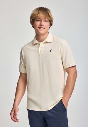 Camisa polo color crema con collar, tapeta de tres botones y mangas cortas. Presenta un pequeño logo negro en el área del pecho izquierdo.