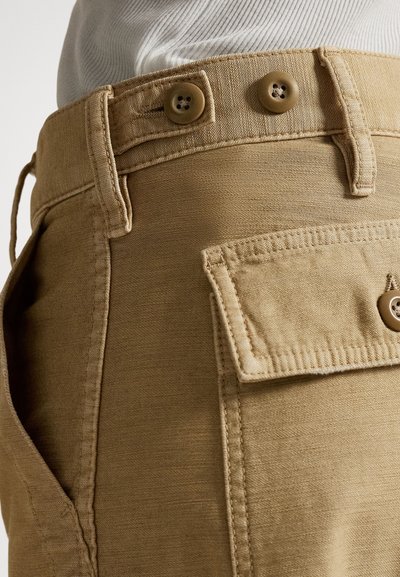 Bež bombažne hlače z gumbnim pasom, stranskimi žepi in zakrpnim žepom na hrbtni strani. Teksturirana tkanina z vidnimi šivi.