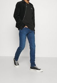 Person som går klädd i svart jacka, svart Puma-t-shirt, blå jeans och svarta Converse-sneakers mot en enfärgad ljus bakgrund.