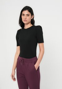 Top nero aderente con maniche corte a sbuffo, abbinato a pantaloni viola scuro con vita a coulisse e tasche laterali. Design minimalista.