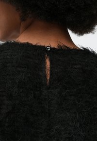 Hátulról látható egy göndör hajú ember, aki egy fekete, bolyhos textúrájú felsőt visel, amelyet a nyak hátsó részén egy fekete gomb zár.