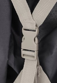 Gros plan d'une boucle en plastique beige portant la marque Hedgren, fixant deux sangles tissées beiges sur un fond de tissu foncé.
