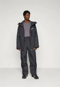 Μαύρο αδιάβροχο μπουφάν με πορτοκαλί λεπτομέρειες και εφαρμοστά παντελόνια, με υφή σχεδίασης και λογότυπα στο στήθος και στο μηρό.