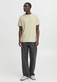 Lys gul flekket t-skjorte med rund hals og brystlomme, kombinert med mørke vide jeans og grå joggesko.