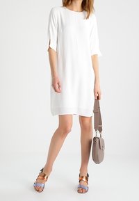 Vestido blanco de manga corta con sutiles aberturas laterales, combinado con sandalias multicolor con detalles dorados, azules y marrones, y un bolso gris.