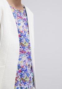Witte, gestructureerde blazer over een top met bloemenprint in blauwe, roze en groene tinten. De blazer heeft een strakke snit met zijzakken.