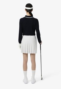 Suéter azul marino de canalé, falda de golf blanca con pliegues, calcetines blancos, zapatillas blancas, sosteniendo un palo de golf. Presenta un diseño deportivo con colores contrastantes.