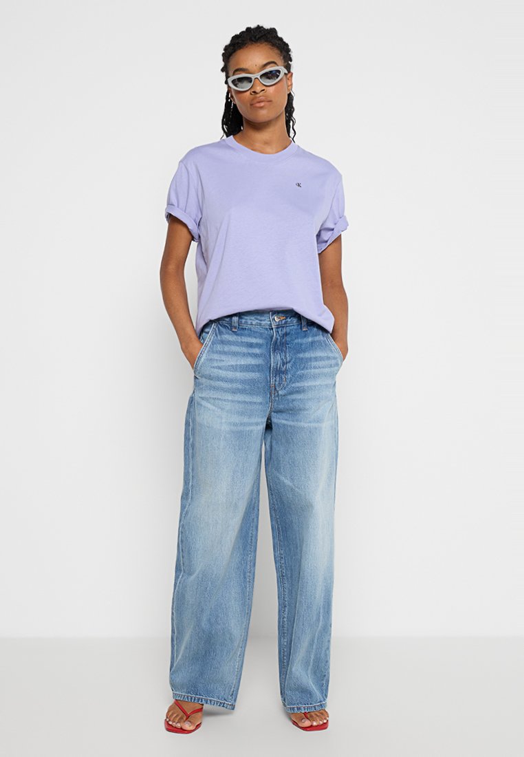 Kobieta w lawendowej koszuli, szerokich niebieskich dżinsach, czerwonych szpilkach i białych okularach przeciwsłonecznych, stojąca z rękami w kieszeniach.