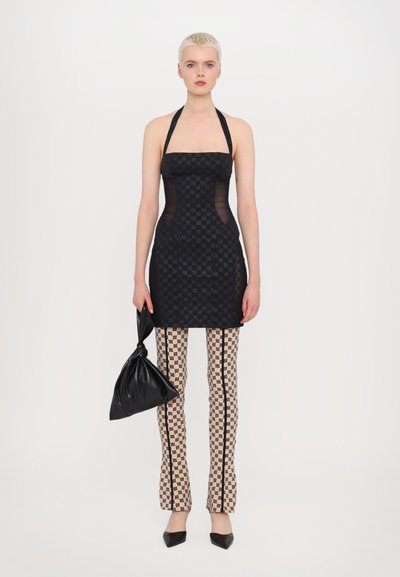 Rochie mini neagră cu decolteu halter, cu panouri laterale transparente, asortată cu pantaloni bej cu un model grafic și o geantă din material negru.