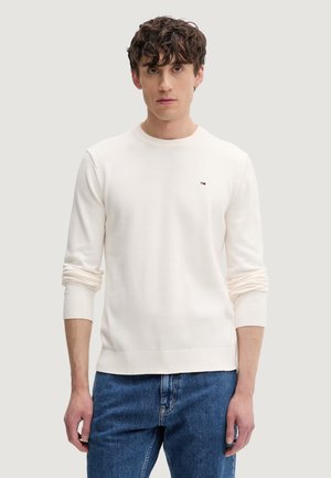 Giovane uomo con capelli ricci che indossa un maglione bianco semplice a maniche lunghe e jeans blu, in piedi davanti a uno sfondo neutro.