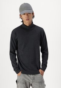 Sort turtleneck sweater med ribbet hals og manchetter, lavet af et blødt stof. Kombineret med en lys grå kasket og afslappede grå bukser.
