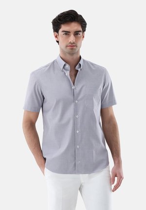 Junger Mann mit dunklem Haar, der ein hellgraues kurzärmeliges Hemd und weiße Hose trägt, steht mit einer Hand in der Tasche.