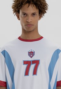 Bala sporta krekls ar sarkanu apkakli un akcentiem, ar ziliem diagonāliem svītrām, logotipu ar zvaigznēm un skaitli 77 treknā sarkanā krāsā.