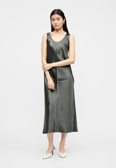 Γυναίκα που στέκεται με σταυρωμένα τα χέρια, φορώντας αμάνικο, γυαλιστερό, γκρι midi φόρεμα και λευκές μυτερές γόβες, σε ένα απλό φόντο.