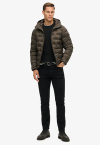 Donkergroene pufferjas met capuchon en een doorstikte structuur. Gemaakt over een zwarte trui, gecombineerd met zwarte jeans en zwarte schoenen.