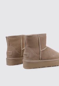 Botas de tobillo de gamuza marrón con suela de goma plana, con costuras cosidas y una etiqueta de marca en el talón. Textura suave, diseño minimalista.