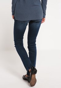 Tmavě modré skinny džíny s hladkou texturou, s zadními kapsami a nízkým pasem, v kombinaci s černými botami na nízkém podpatku.