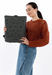 Mustreeritud musta geomeetrilise mustriga seljakott, millel on tekstureeritud pind, ülemine käepide ja sile alus, mille hoiab käes isik vabaaja riietuses.