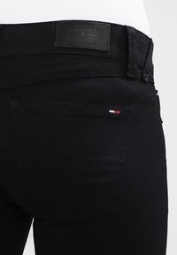 Los pantalones vaqueros negros presentan una textura suave con una etiqueta en la cinturilla y una pequeña etiqueta de logotipo tricolor en el bolsillo trasero. Diseño simple y corte ajustado.
