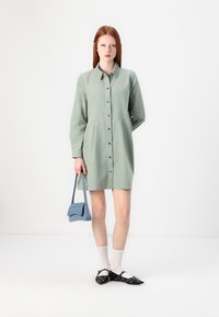 Abito camicia verde chiaro con chiusura a bottoni, maniche lunghe, una vestibilità rilassata e una singola tasca sul petto. Abbinato a una borsa blu e scarpe nere.