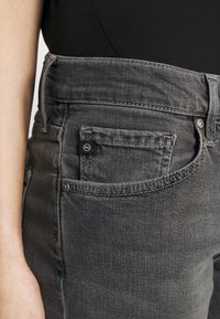 Jeans en denim gris avec une taille mi-haute, des détails de poche avant et une petite étiquette de marque sur la poche arrière. Texture lisse.