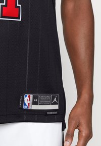 Juodas krepšinio marškinėliai su raudonu numeriu "1". Fiksuojama tinklelio tekstūra ir baltos detalės. Etiketėje nurodyta "NBA Authentic" ir "Jordan". Dydis 44.