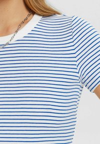 Biała koszula z krótkim rękawem w cienkie poziome niebieskie paski, noszona z srebrnym naszyjnikiem, odsłaniająca okolice szyi i ramion.