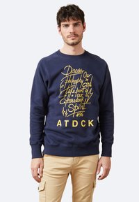 Sudadera azul marino hecha de algodón texturizado con texto gráfico amarillo en la parte delantera, que presenta un cuello redondo y puños y dobladillo acanalados.
