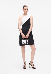 Fekete ruha egy fehér vállpánttal, minimalista design. A modell egy kis fekete-fehér táskával a kezében, fekete pántos magassarkú cipőt visel.