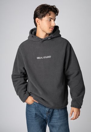 Gray fleece hoodie with a soft texture, featuring a front logo "SBLVL STUDIO" in white. Paired with blue denim jeans.