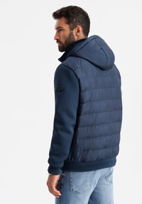 Mörkblå pufferjacka med huva, som har en slät yta, ribbade mudd och en logodetalj på ärmen. Stilig och varm design.