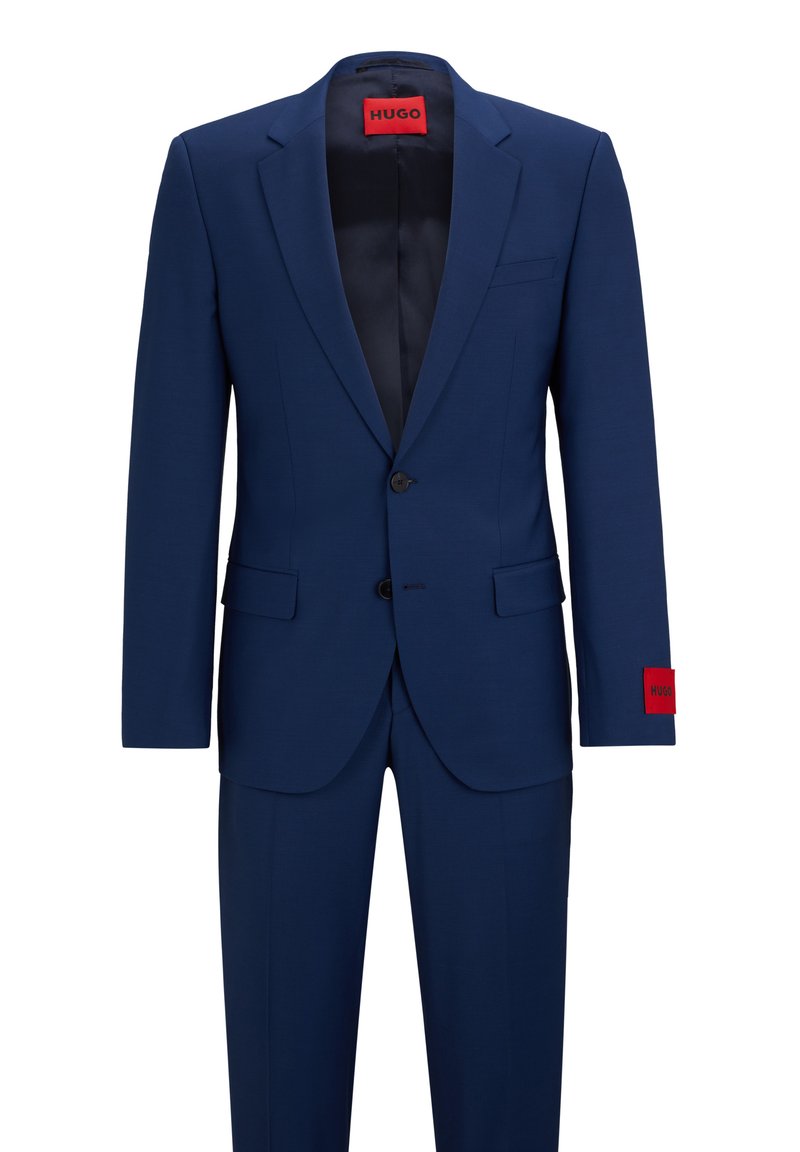 Marineblauer Anzug mit einem einreihigen Blazer, zwei Knöpfen und geregelten Revers. Rotes Etikett am Ärmel; glatte Textur und taillierter Schnitt.