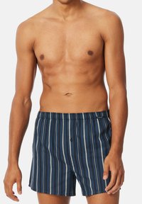 Boxer da uomo a righe nei toni del blu e del navy, con righe verticali, una vita elastica e un dettaglio di un solo bottone frontale.