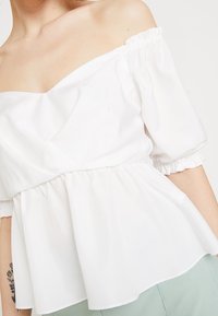 Bílá blůza bez ramínek s krátkými nabíranými rukávy a elastickými manžetami. Má stažený pas a hladkou strukturu.