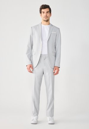 Traje gris claro con solapas de muesca, dos botones y bolsillos; llevado sobre una camisa blanca. Acompañado de zapatillas blancas, con una textura suave.