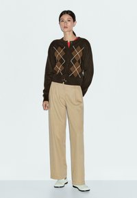 Brauner Argyle-Cardigan mit cremefarbenen und braunen Mustern, Knopfverschluss, kombiniert mit leicht beigefarbenen, weit geschnittenen Hosen und weißen Schuhen.