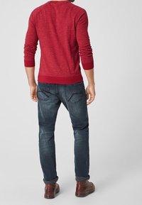 Maglione rosso a maglia con polsini e orlo a costine, abbinato a jeans blu scuro con una texture sbiadita e orli arrotolati, indossato con scarpe marroni.